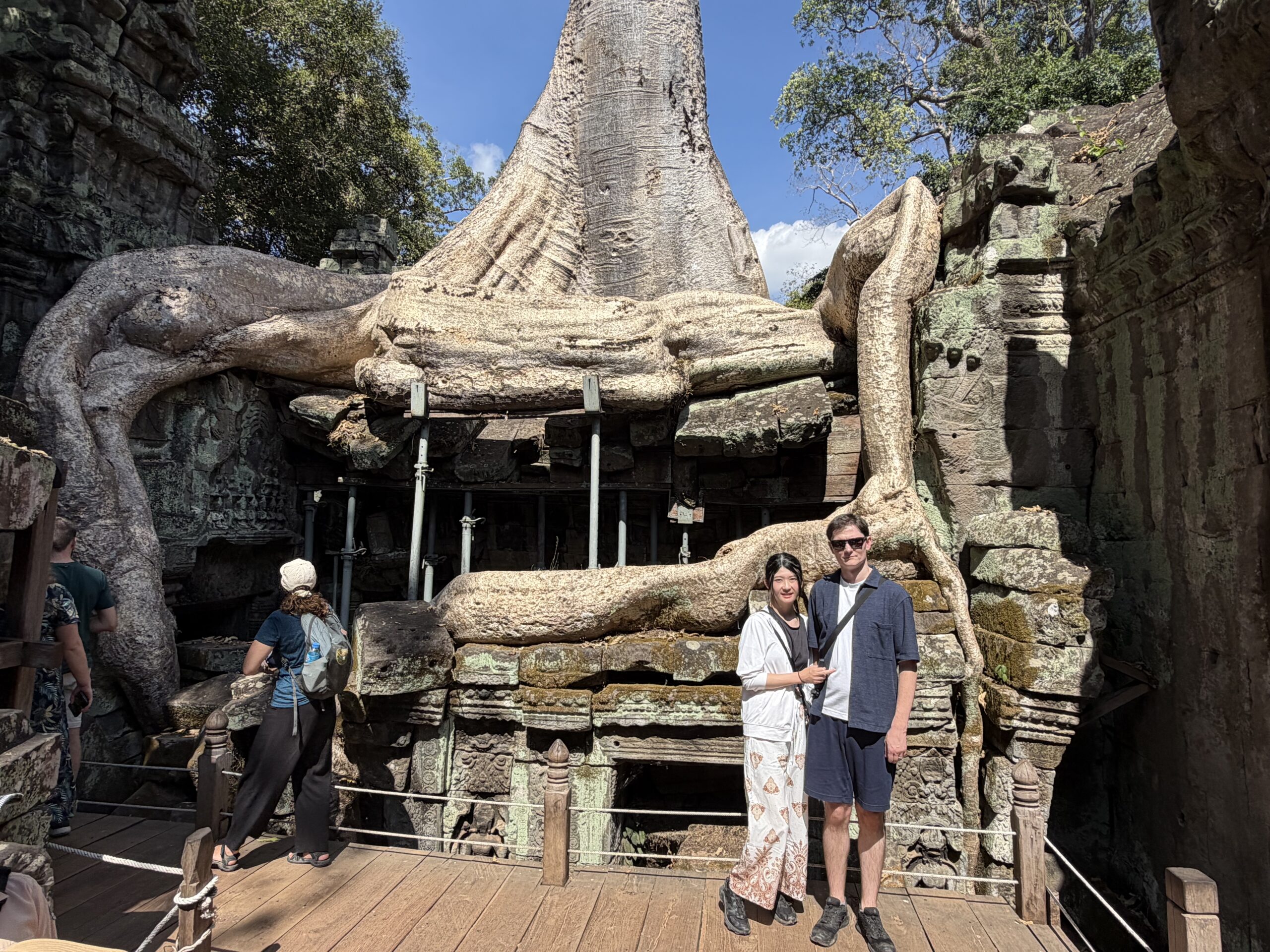Two people standing before massive tree roots at the Ta Prohm ruins